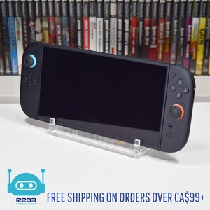 May include: A black handheld gaming console with a black screen, blue and orange accents, and a clear acrylic stand. The console is displayed in front of a shelf of video game cases.