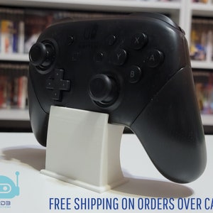 May include: A black Nintendo Switch Pro Controller is displayed on a white plastic stand. The controller has a black and white design with buttons labeled X, Y, A, and B. The stand is a simple design with a rectangular base and a raised platform to hold the controller.