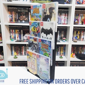 May include: A black stand with a clear plastic insert holds a collection of Nintendo Switch game cartridges. The cartridges feature various game titles, including "Animal Crossing", "Batman", "Bubble Bobble", "Cavestory", and "Toad Treasure Tracker". The stand is displayed on a white shelf with other video game cartridges.