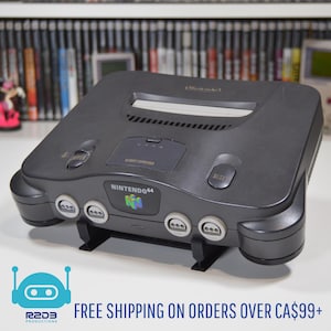 Puede incluir: Una consola de videojuegos Nintendo 64 gris oscuro con el logotipo de Nintendo 64 en la parte delantera. La consola tiene cuatro puertos de controlador y un botón de reinicio. La consola está sobre un soporte negro.