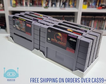 Puede incluir: Una pila de ocho cartuchos de juego de Super Nintendo grises, incluyendo Mortal Kombat, en un estante blanco. Los cartuchos están dispuestos verticalmente, con el cartucho de Mortal Kombat en la parte inferior. El texto "AKlaim" y "MORTAL KOMBAT" es visible en el cartucho de Mortal Kombat.