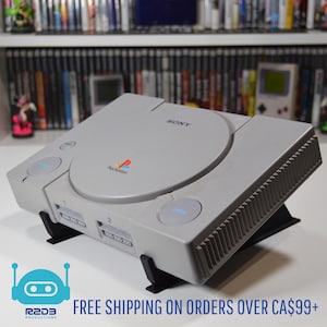 May include: A gray Sony PlayStation 1 video game console, with the PlayStation logo and the word "SONY" visible. The console is on a black stand. The console has two controller ports and a disc tray. The background shows a shelf with video games.