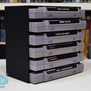 May include: A black storage rack holding six grey video game cartridges stacked horizontally. The labels read Disney's Goof Troop, Barbie Super Model, Super Star Wars, and Pitfall The Mayan Adventure. The rack has a solid black side panel.