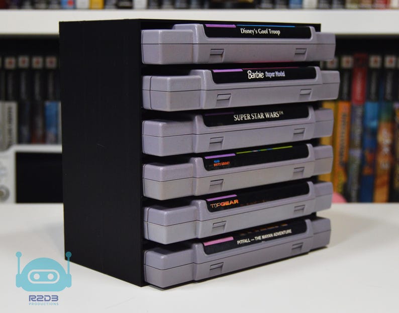 May include: A black storage rack holding six grey video game cartridges stacked horizontally. The labels read Disney's Goof Troop, Barbie Super Model, Super Star Wars, and Pitfall The Mayan Adventure. The rack has a solid black side panel.