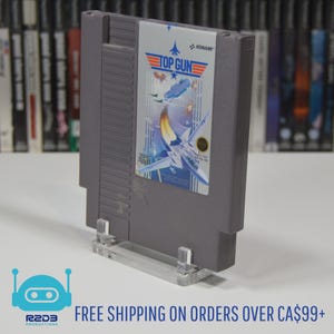 Puede incluir: Un cartucho de juego gris de Nintendo Entertainment System (NES) con una etiqueta que muestra un avión de combate azul y blanco sobre un fondo de cielo azul. La etiqueta dice "Top Gun" y "Konami".