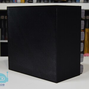 May include: A black rectangular storage unit designed to hold video game cartridges. The unit features a solid black exterior side panel and a series of grey video game cartridges stacked vertically along the right side of the storage structure.