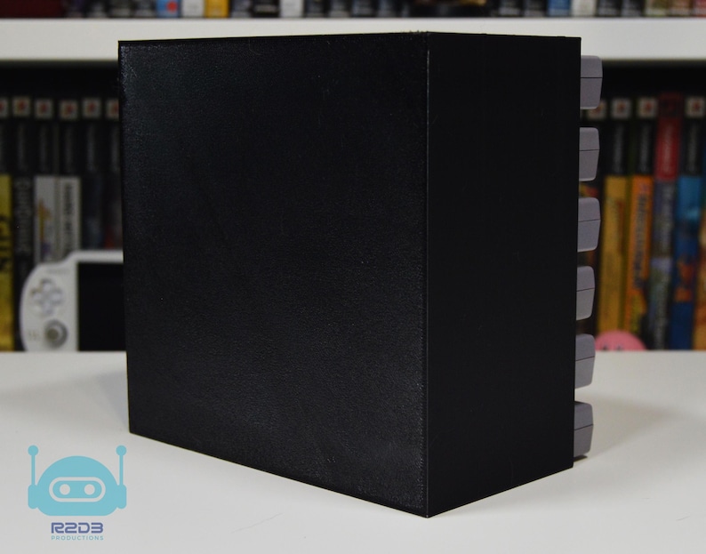 May include: A black rectangular storage unit designed to hold video game cartridges. The unit features a solid black exterior side panel and a series of grey video game cartridges stacked vertically along the right side of the storage structure.