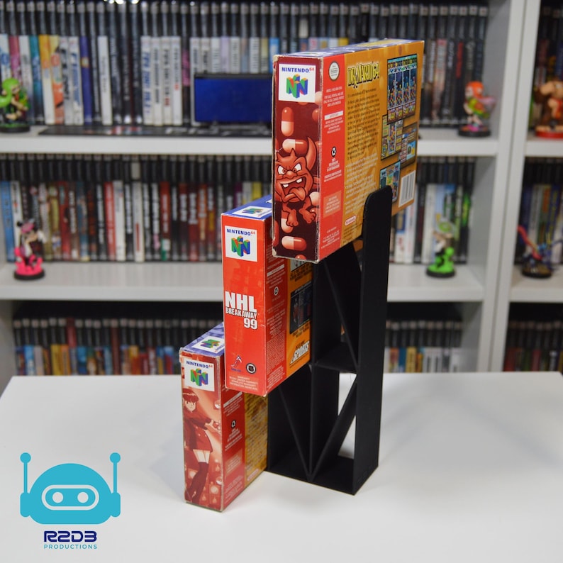 May include: A black plastic stand with three levels for displaying Nintendo 64 game cartridges. The stand is holding three game cartridges: 'NHL Breakaway '99', 'Imps', and 'Kirby's Avalanche'.
