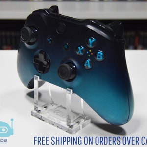 May include: A black and teal gradient video game controller on a clear acrylic stand. The controller has colored buttons and control sticks. The image also includes the text "FREE SHIPPING ON ORDERS OVER CA$99+".