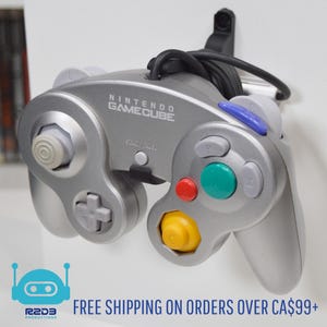 May include: A silver Nintendo GameCube controller with a black cord attached. The controller has a variety of buttons, including a yellow, red, green, and blue button. The controller is mounted on a white wall.