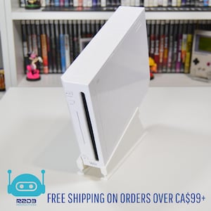 Peut inclure: Une console Nintendo Wii blanche, posée sur un support blanc. La console a un design rectangulaire avec une fente noire verticale. Le mot "Wii" est visible à l'avant. En arrière-plan, une étagère avec des jeux vidéo.