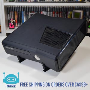 Op de afbeelding: Een zwarte Xbox 360 console, op een zwarte standaard. De console heeft een schijflade, aan/uit-knop en ventilatieroosters. De tekst "XBOX 360" is zichtbaar. De afbeelding bevat ook de tekst "R2D3 Productions".