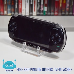 Könnte beinhalten: Schwarze Sony PSP-Handheld-Spielkonsole auf einem transparenten Acryl-Ständer. Die Konsole hat einen Bildschirm, Tasten und einen Joystick. Der Text "PSP" ist auf dem Ständer sichtbar. Im Hintergrund ein Regal mit bunten Spielhüllen.