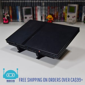 May include: A black PlayStation 2 console on a black stand. The console is rectangular with a central groove and the PlayStation logo. The image has a white background with a shelf of books and game consoles in the background.