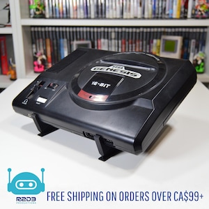 May include: A black Sega Genesis 16-bit video game console. The console has the Sega Genesis logo and "16-BIT" text on the top. The console is on a white surface. The image also includes the R2D3 Productions logo.