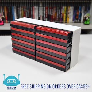 May include: A stack of black and red game cartridges in a white holder. The cartridges have red labels and are stacked neatly. The holder is white and has a rectangular shape. The background shows a shelf with other games.