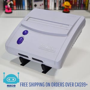 May include: A light grey Super Nintendo Entertainment System console with two purple buttons. The console has the words "Super Nintendo Entertainment System" printed on the front. The console is on a black stand.