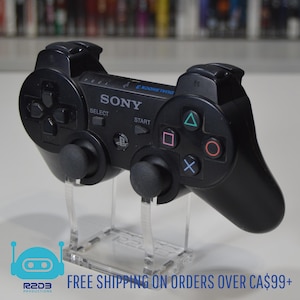 Op de afbeelding: Een zwarte Sony DualShock 3 controller voor de Playstation 3 console. De controller staat op een heldere acryl standaard. De controller heeft een zwart-wit ontwerp met de woorden "DUALSHOCK 3" en "SONY" erop gedrukt.