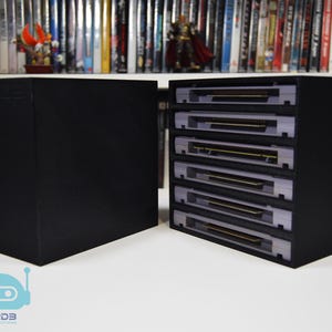May include: A black rectangular storage unit with five horizontal slots, each containing a grey video game cartridge. The unit is positioned next to a solid black side panel. A light blue robot icon with the text R2D3 PRODUCTIONS is visible.