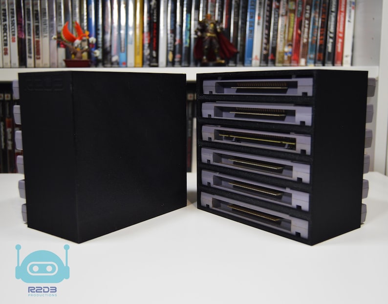 May include: A black rectangular storage unit with five horizontal slots, each containing a grey video game cartridge. The unit is positioned next to a solid black side panel. A light blue robot icon with the text R2D3 PRODUCTIONS is visible.