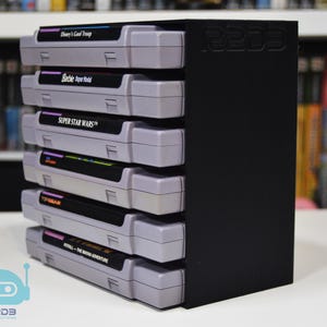 May include: A black vertical storage rack holding six gray video game cartridges stacked on their sides. The rack features the embossed text R2D3 on the side panel. The cartridges include titles such as Disney's Goof Troop, Barbie Super Model, and Super Star Wars.