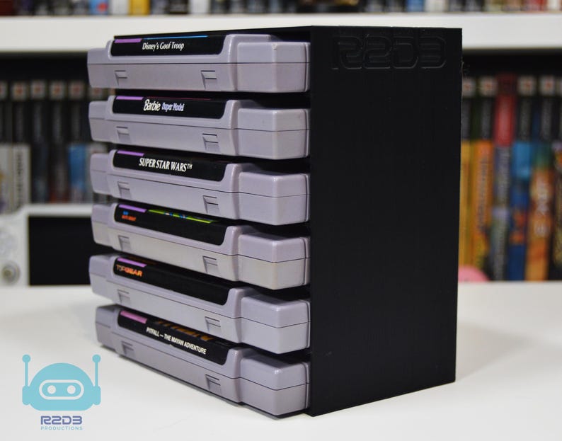 May include: A black vertical storage rack holding six gray video game cartridges stacked on their sides. The rack features the embossed text R2D3 on the side panel. The cartridges include titles such as Disney's Goof Troop, Barbie Super Model, and Super Star Wars.