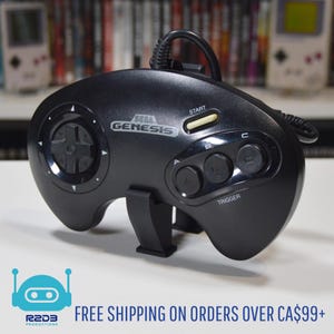 May include: Black Sega Genesis controller with a directional pad, start button, and three action buttons. The controller has the Sega Genesis logo and the word "TRIGGER" printed on it. The controller is on a white surface.