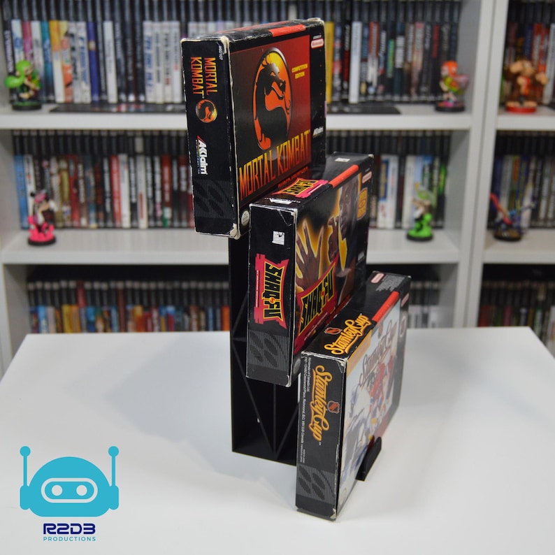 May include: Three vintage Super Nintendo game cartridges stacked on a black plastic stand. The cartridges are for the games Mortal Kombat, Shaq Fu, and Street Fighter II: Champion Edition. The cartridges are black with colorful labels.