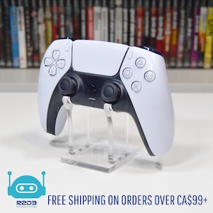 May include: A white and black PlayStation 5 controller on a clear acrylic stand. The controller has a black center section with buttons and joysticks. The stand is square and has a flat base. The controller is in front of a shelf of video games.