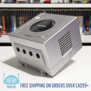 Może przedstawiać: Srebrna konsola Nintendo GameCube. Konsola ma okrągłą szufladę na płyty i kilka portów z przodu. Logo GameCube jest widoczne na górze. Konsola stoi na białej powierzchni.