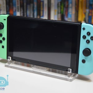 May include: A black Nintendo Switch console with a mint green and light blue Joy-Con controller. The console is on a clear acrylic stand.