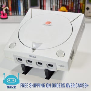 Puede incluir: Consola de juegos Sega Dreamcast blanca con el logotipo de Dreamcast en la parte superior. La consola tiene una bandeja de disco, puertos de controlador y un botón "Open". La consola está soportada por un soporte negro. El texto "R2D3 Productions" y "FREE SHIPPING ON ORDERS OVER CA$99+" están en la parte inferior.