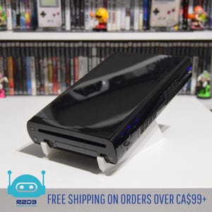Puede incluir: Una consola de videojuegos negra sobre un soporte blanco. La consola es brillante y rectangular, con una ranura para unidad de disco. El soporte es blanco e inclinado. El fondo incluye una estantería de cajas de videojuegos.