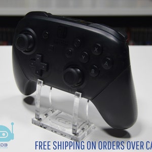 May include: A black Nintendo Switch Joy-Con controller on a clear acrylic stand. The controller has the Nintendo Switch logo and buttons labelled X, Y, A, and B.
