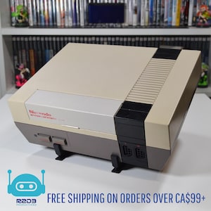 Op de afbeelding: Een beige en grijze Nintendo Entertainment System (NES) console. De console heeft een rechthoekig ontwerp met een sleuf voor spelcartridges en controllerpoorten. Het Nintendo-logo is zichtbaar aan de voorkant. De console wordt weergegeven op een wit oppervlak.