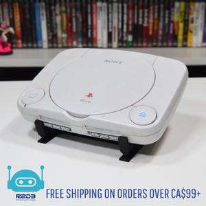 May include: A white Sony PSone video game console with a black stand. The console has a disc tray, power and reset buttons, and memory card slots. The PSone logo is visible.