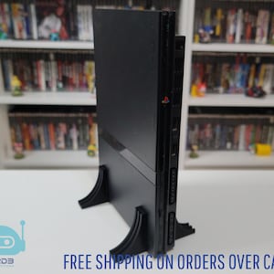 May include: A black PlayStation 2 console standing upright on a black stand. The console is positioned vertically, showcasing its sleek design. The text "FREE SHIPPING ON ORDERS OVER CA$99+" is visible at the bottom.