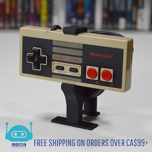 Op de afbeelding: Een zwarte standaard met een beige Nintendo-controller die eraan is bevestigd. De controller heeft een zwarte directionele pad, twee zwarte knoppen en twee rode knoppen met de labels "B" en "A". De controller heeft de tekst "SELECT START" en "Nintendo" erop gedrukt.