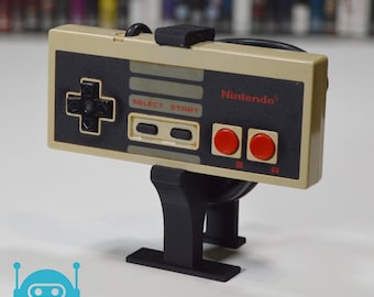 May include: A black stand with a beige Nintendo controller attached to it. The controller has a black directional pad, two black buttons, and two red buttons labeled "B" and "A". The controller has the text "SELECT START" and "Nintendo" printed on it.