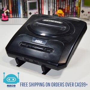May include: Black Sega Genesis video game console. The console has the word "SEGA" printed on the front. The console is sitting on a black stand. The image also includes the text "FREE SHIPPING ON ORDERS OVER CA$99+".