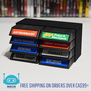 May include: Black game cartridge storage unit holding eight game cartridges. Cartridge labels include "ASTROSMASH", "AUTO RACING", "BOXING", "DUNGEONS & DRAGONS, TREASURE of TARMIN", "Poker & Blackjack", "NIGHT STALKER", "PINBALL", and "SKIING".