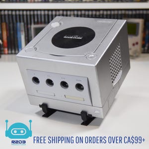 Może przedstawiać: Srebrna konsola Nintendo GameCube z logo GameCube na górze. Konsola ma czarną tacę na dyski i kilka portów z przodu. Konsola stoi na czarnym stojaku. Obraz zawiera również logo R2D3 Productions.
