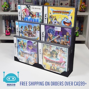 May include: A black display stand with multiple tiers, showcasing various Nintendo DS game cases. The game cases display vibrant artwork and titles such as "Dragon Quest" and "Rocket Slime." The stand is on a white surface, with a bookshelf visible.