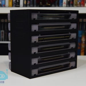 May include: A black rectangular storage rack designed to hold six grey video game cartridges in a vertical stack. The unit features six horizontal slots, each containing a grey cartridge, with the connector pins visible at the back of each slot.