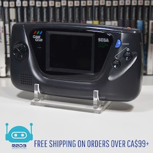Könnte beinhalten: Eine schwarze Sega Game Gear Handheld-Videospielkonsole mit einem klaren Acryl-Ständer. Die Konsole hat einen Farbbildschirm, einen Startknopf und den Text "GAME GEAR" und "SEGA" darauf gedruckt.
