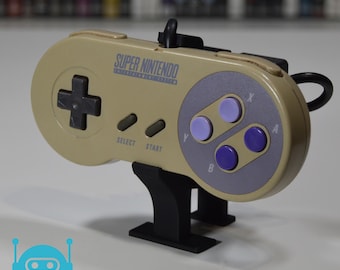 May include: A Super Nintendo controller in a beige color, with purple and black buttons, and the words "Super Nintendo" printed on it. The controller is mounted on a black stand. The controller has a directional pad, select, start, X, Y, A, and B buttons.