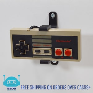 Puede incluir: Un soporte de pared negro para un mando de control de Nintendo Entertainment System. El mando es beige con botones negros y botones A y B rojos. El texto "Nintendo" está impreso en el mando.