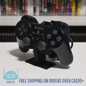 May include: A black Sony PlayStation controller is displayed on a black stand. The controller has directional buttons, analog sticks, and action buttons with colored symbols. The text "SONY" and "PlayStation" are visible on the controller.