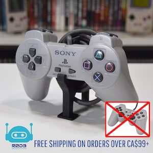 Peut inclure: Une manette grise Sony PlayStation avec des boutons noirs et un câble noir. La manette est sur un support noir. Les textes "SONY" et "PlayStation" sont visibles sur la manette.