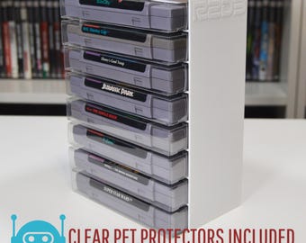 Puede incluir: Soporte de acrílico blanco con fundas de plástico transparente para guardar cartuchos de juegos de Super Nintendo. El soporte tiene capacidad para ocho cartuchos y lleva el texto "R2D3 Productions" en el lateral. Los cartuchos incluyen títulos como "SimCity", "Jurassic Park" y "Super Star Wars".